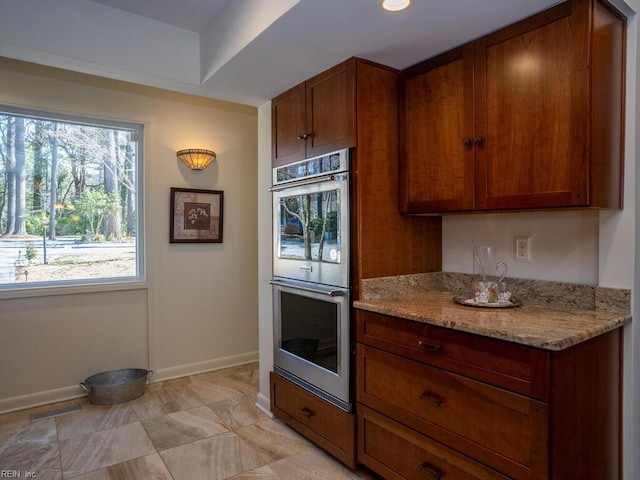 kitchen featuring light stone counters, visible vents, brown cabinetry, stainless steel double oven, and baseboards