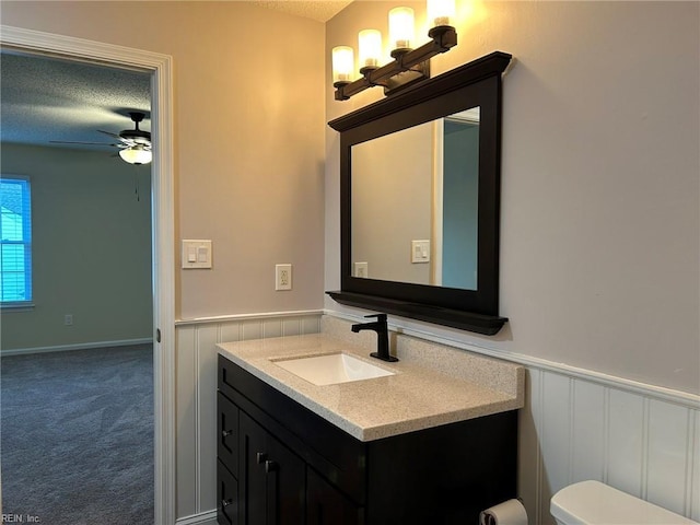 half bath with a ceiling fan, toilet, a wainscoted wall, a textured ceiling, and vanity