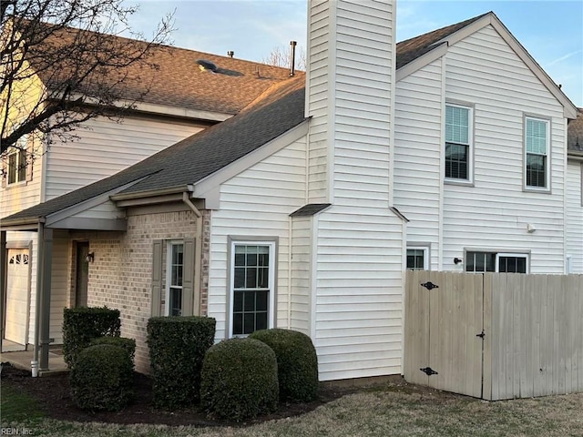 view of side of property featuring a garage, brick siding, a shingled roof, fence, and a chimney