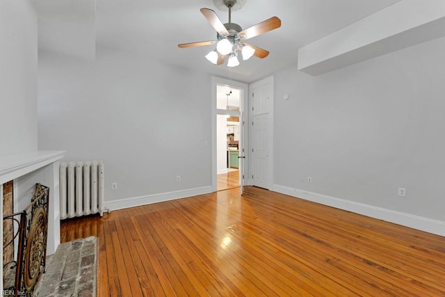 unfurnished room featuring ceiling fan, baseboards, a brick fireplace, radiator, and hardwood / wood-style floors