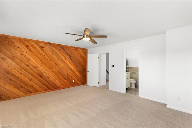 unfurnished bedroom with light carpet, wooden walls, baseboards, connected bathroom, and ceiling fan