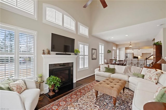 living room with dark wood-style flooring, a towering ceiling, a ceiling fan, a glass covered fireplace, and baseboards