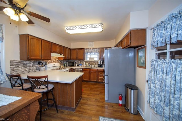 kitchen featuring appliances with stainless steel finishes, a peninsula, light countertops, under cabinet range hood, and backsplash