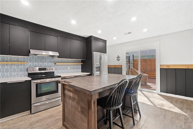 kitchen with stainless steel appliances, backsplash, dark cabinets, under cabinet range hood, and a kitchen breakfast bar