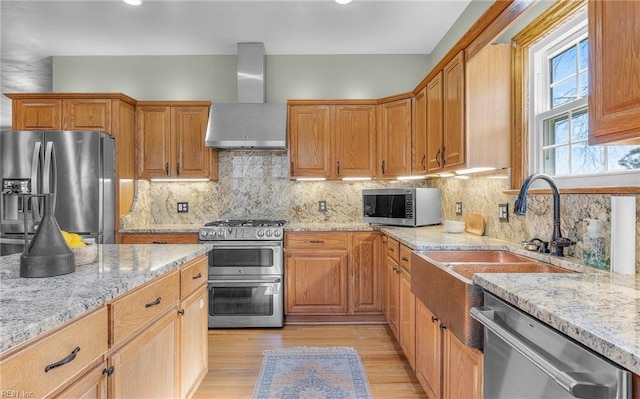 kitchen with light wood-style flooring, a sink, wall chimney range hood, appliances with stainless steel finishes, and light stone countertops