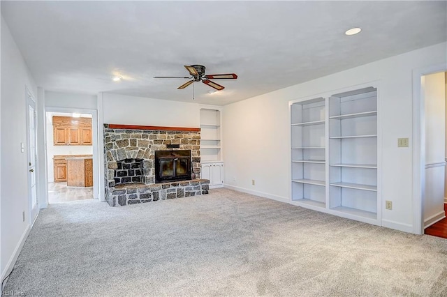 unfurnished living room featuring built in shelves, a ceiling fan, carpet flooring, a stone fireplace, and baseboards