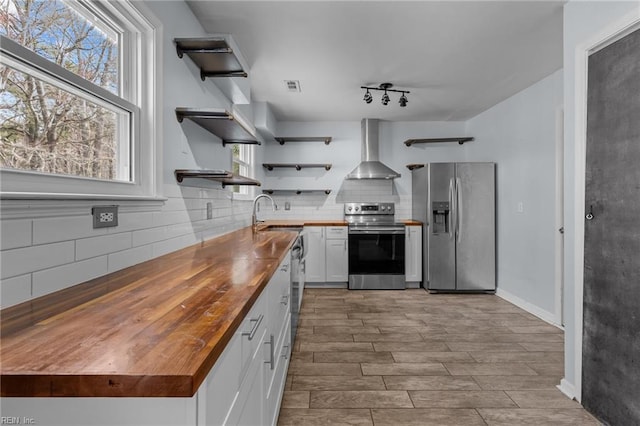 kitchen featuring open shelves, stainless steel appliances, a sink, wood counters, and wall chimney exhaust hood