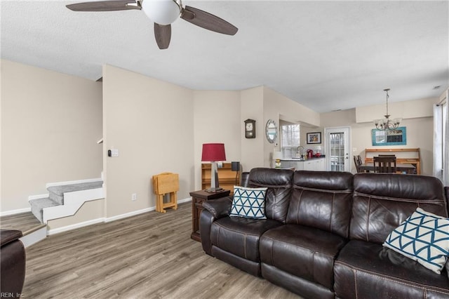 living room featuring stairs, baseboards, wood finished floors, and ceiling fan with notable chandelier