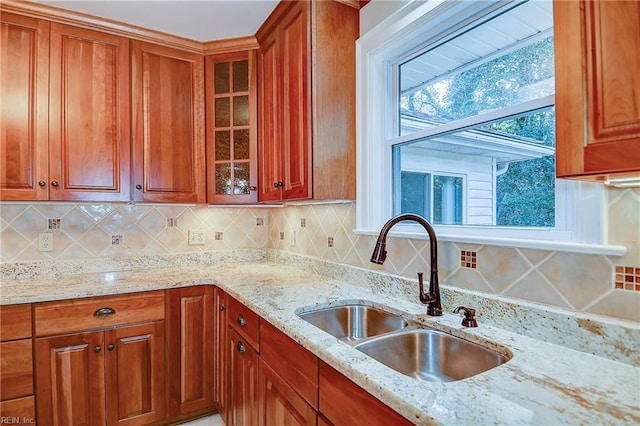 kitchen featuring light stone counters, a sink, decorative backsplash, brown cabinetry, and glass insert cabinets