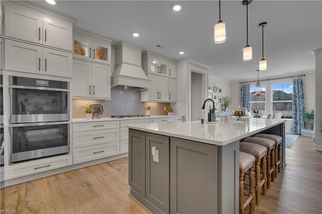 kitchen with premium range hood, light wood-style floors, appliances with stainless steel finishes, tasteful backsplash, and a kitchen bar