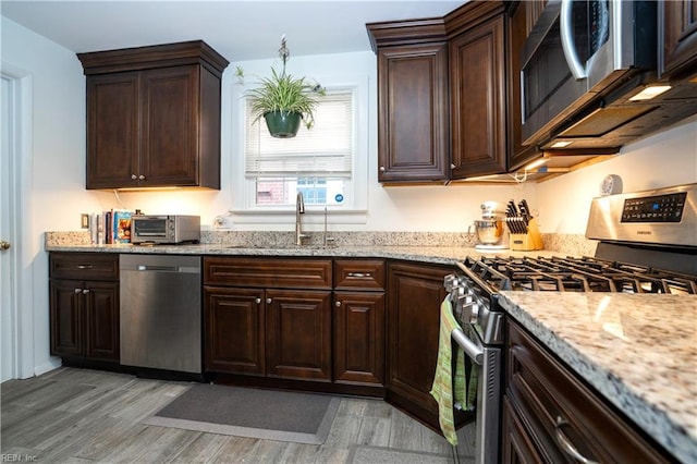 kitchen featuring light wood finished floors, stainless steel appliances, a sink, dark brown cabinetry, and light stone countertops