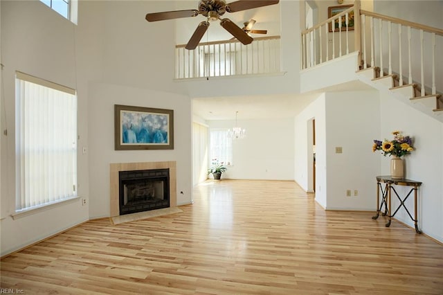 unfurnished living room with a tile fireplace, ceiling fan with notable chandelier, a towering ceiling, light wood-style floors, and stairway