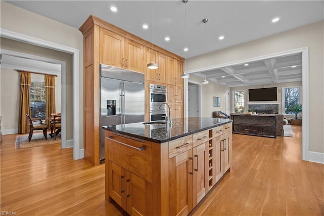 kitchen featuring a center island with sink, coffered ceiling, a sink, stainless steel appliances, and light wood-type flooring