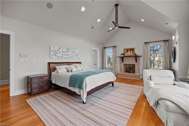 bedroom featuring a fireplace with raised hearth, baseboards, light wood-style flooring, high vaulted ceiling, and a ceiling fan