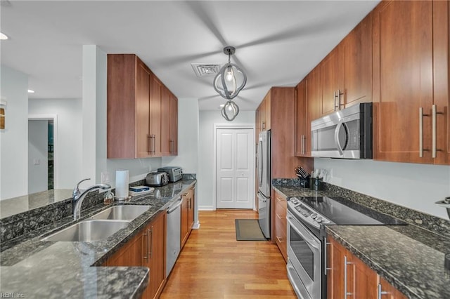 kitchen with brown cabinets, appliances with stainless steel finishes, a sink, dark stone countertops, and light wood-type flooring