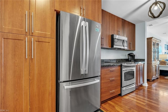 kitchen with light wood finished floors, appliances with stainless steel finishes, dark stone counters, and brown cabinetry