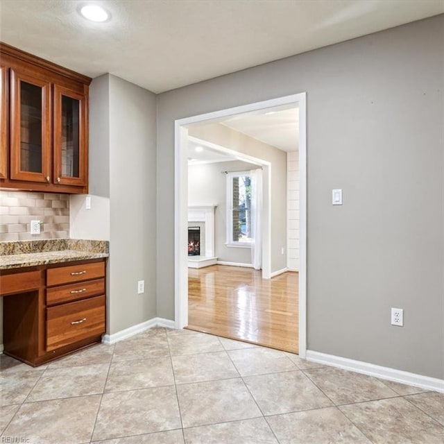 kitchen featuring brown cabinets, light tile patterned floors, glass insert cabinets, a lit fireplace, and baseboards