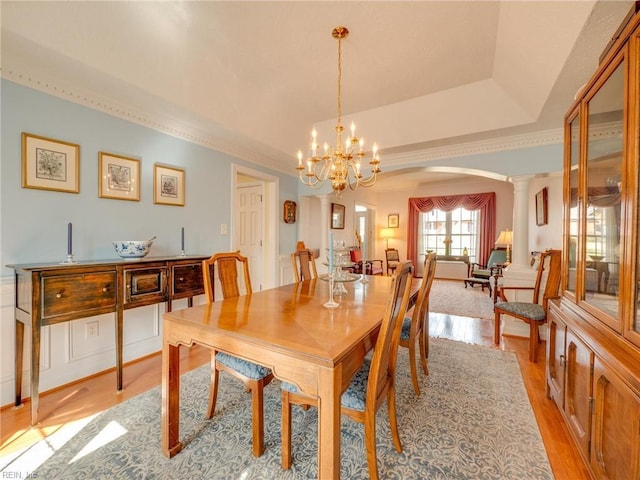 dining space featuring arched walkways, light wood-style flooring, an inviting chandelier, a tray ceiling, and ornate columns