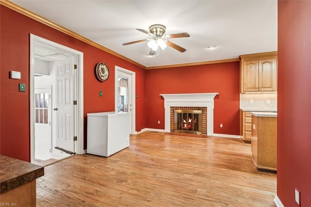 unfurnished living room with light wood finished floors, baseboards, a ceiling fan, ornamental molding, and a brick fireplace