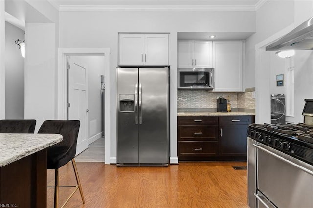 kitchen with ornamental molding, appliances with stainless steel finishes, light wood-type flooring, wall chimney range hood, and a kitchen bar