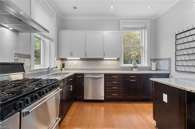 kitchen featuring light stone counters, stainless steel appliances, crown molding, wall chimney range hood, and a sink