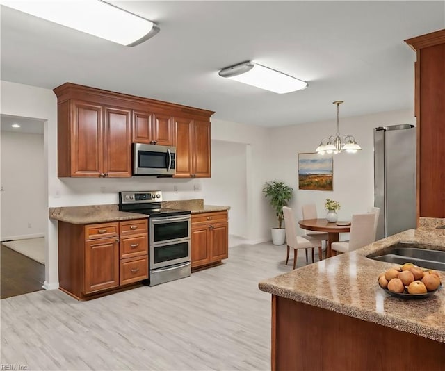 kitchen featuring light stone countertops, light wood-type flooring, brown cabinets, appliances with stainless steel finishes, and hanging light fixtures