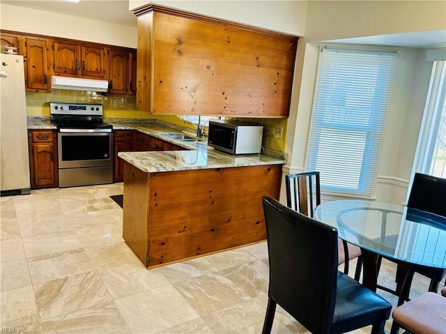 kitchen with light stone counters, under cabinet range hood, a peninsula, appliances with stainless steel finishes, and backsplash