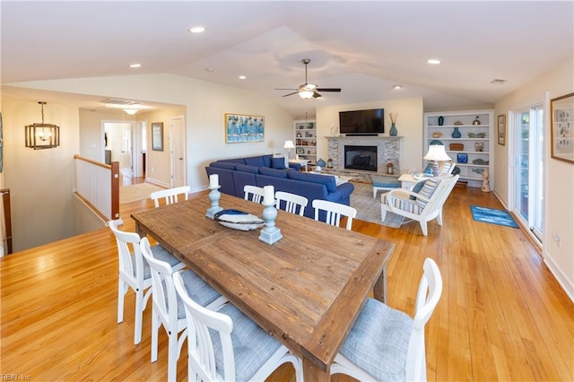 dining space featuring built in shelves, light wood finished floors, a ceiling fan, vaulted ceiling, and a stone fireplace