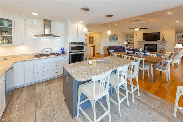 kitchen with stainless steel appliances, a fireplace with raised hearth, open floor plan, white cabinets, and wall chimney exhaust hood