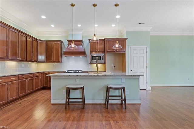 kitchen featuring custom exhaust hood, tasteful backsplash, stainless steel microwave, a kitchen island with sink, and wood finished floors