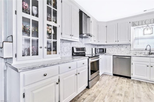 kitchen with stainless steel appliances, a sink, white cabinetry, wall chimney range hood, and glass insert cabinets