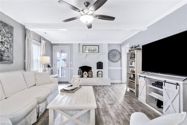 living area with ceiling fan, wood finished floors, ornamental molding, a brick fireplace, and beamed ceiling