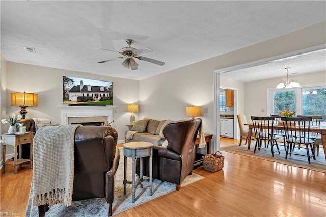 living room with a textured ceiling, ceiling fan with notable chandelier, a fireplace, visible vents, and light wood finished floors