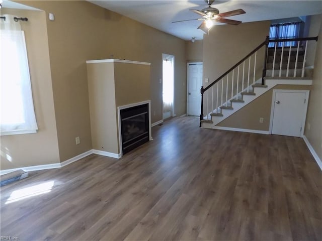 unfurnished living room with a glass covered fireplace, ceiling fan, wood finished floors, baseboards, and stairs