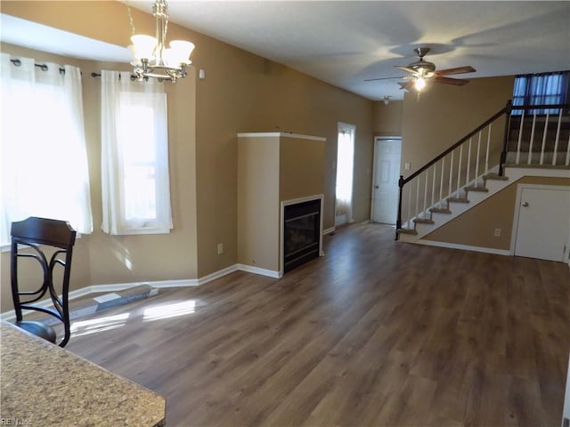 unfurnished living room featuring stairs, a glass covered fireplace, wood finished floors, baseboards, and ceiling fan with notable chandelier