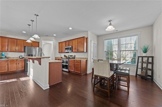 kitchen with brown cabinets, hanging light fixtures, appliances with stainless steel finishes, dark wood-type flooring, and light stone countertops