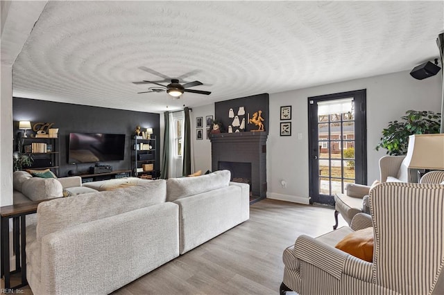 living area with a brick fireplace, ceiling fan, a textured ceiling, light wood-type flooring, and baseboards