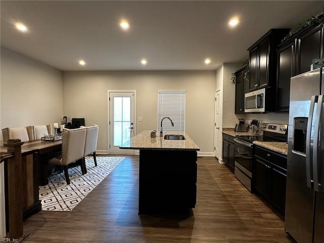 kitchen featuring a kitchen island with sink, dark cabinets, dark wood-style flooring, a sink, and appliances with stainless steel finishes