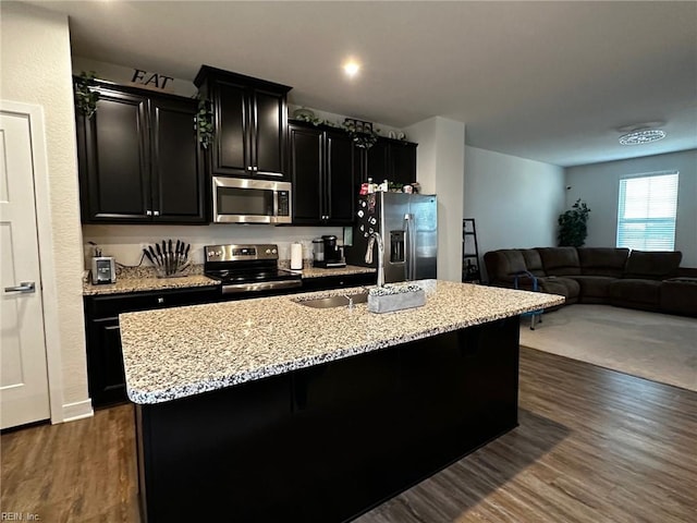 kitchen with an island with sink, dark wood-style floors, appliances with stainless steel finishes, dark cabinets, and a sink