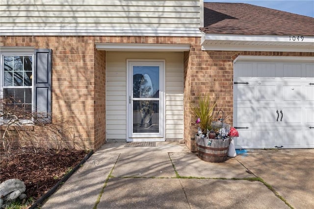 view of exterior entry with brick siding, an attached garage, driveway, and a shingled roof