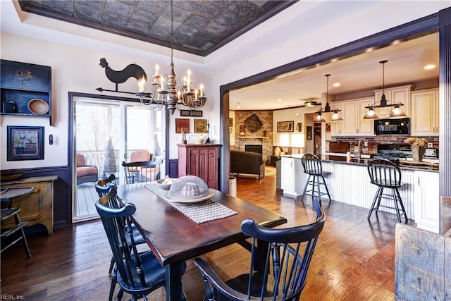 dining space featuring a raised ceiling, a wainscoted wall, an ornate ceiling, dark wood-style flooring, and a stone fireplace