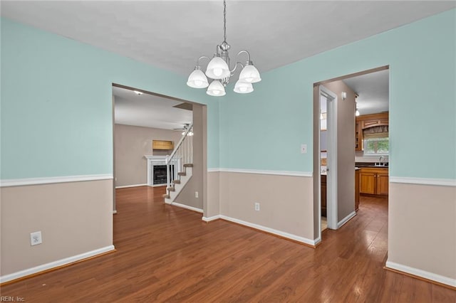 unfurnished dining area featuring a fireplace, a sink, wood finished floors, baseboards, and ceiling fan with notable chandelier