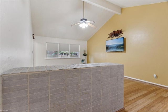 interior space with a ceiling fan, wood finished floors, baseboards, vaulted ceiling with beams, and tile counters