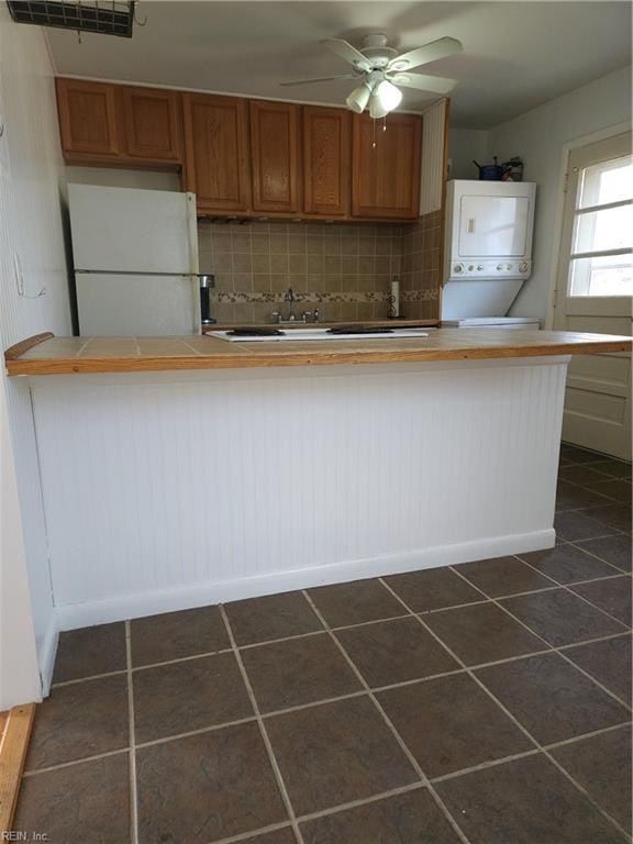 kitchen with stacked washer / drying machine, decorative backsplash, brown cabinetry, freestanding refrigerator, and dark tile patterned flooring