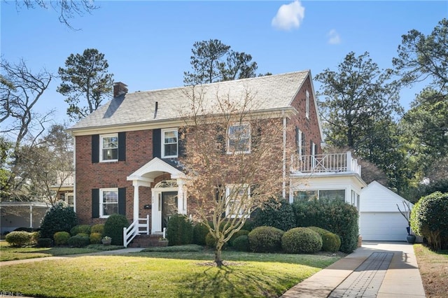 view of front of property with brick siding, a front yard, a chimney, a balcony, and an outbuilding