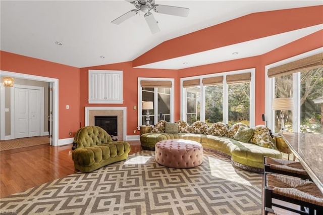 living area featuring baseboards, vaulted ceiling, wood finished floors, a glass covered fireplace, and a ceiling fan