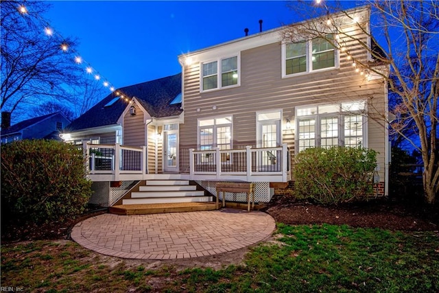 back of house at twilight featuring a wooden deck, a patio, and roof with shingles