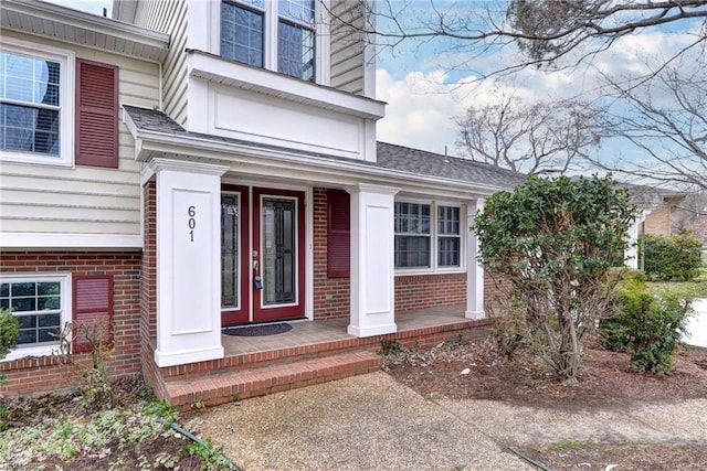 view of exterior entry with covered porch, a shingled roof, and brick siding