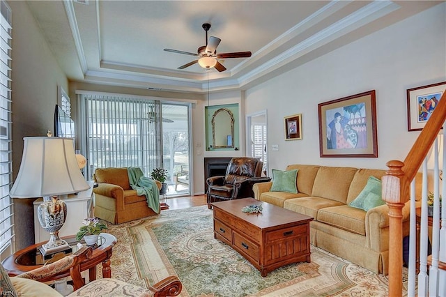 living area with ceiling fan, light wood-style flooring, a fireplace, ornamental molding, and a tray ceiling