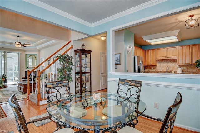 dining space featuring a ceiling fan, light wood-style floors, stairs, ornamental molding, and a raised ceiling